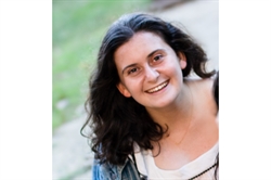 [ai] A woman with long, curly hair smiles while seated outdoors, wearing a light top and a denim jacket. The background features blurred greenery, giving a natural setting.